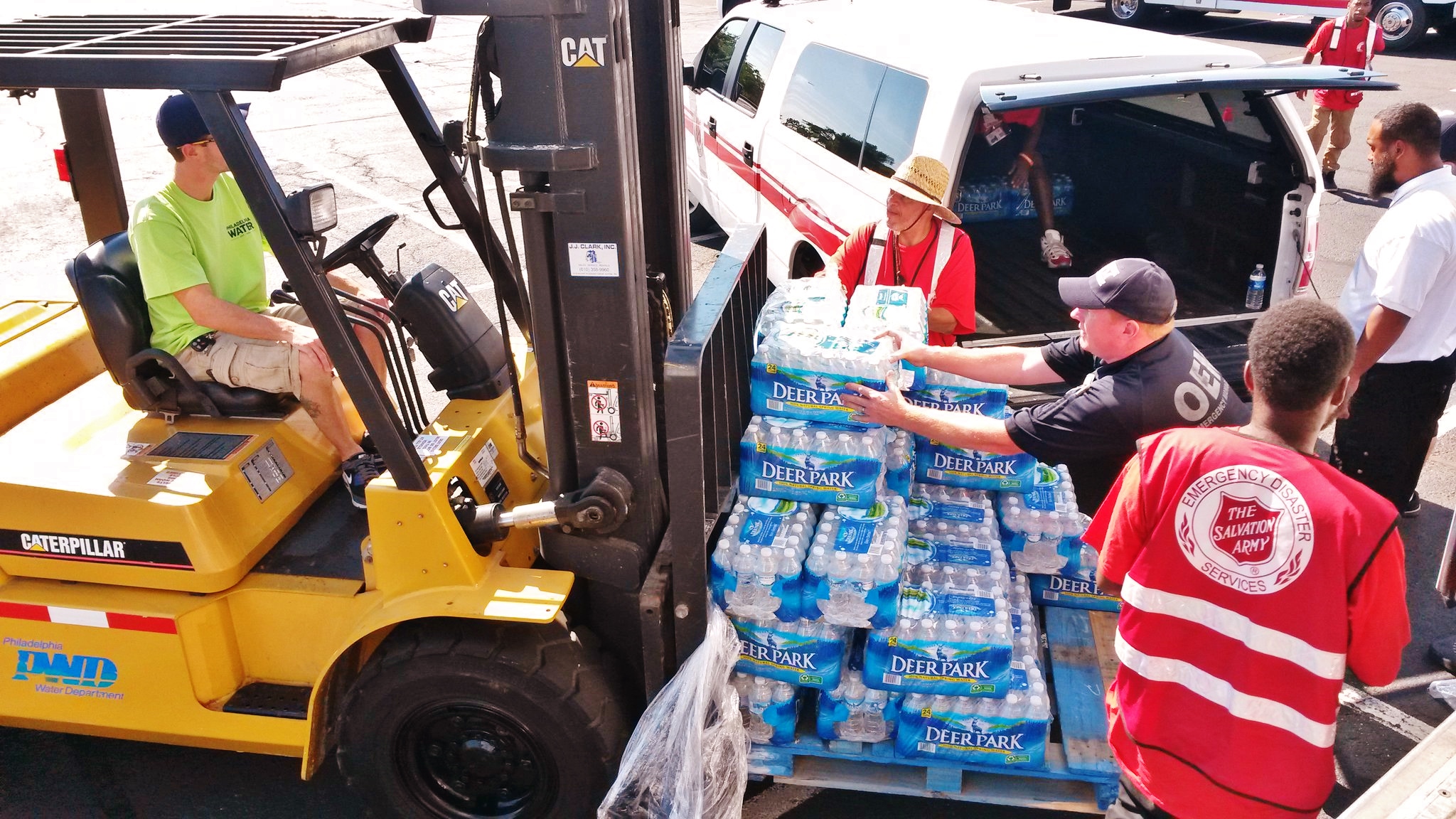 Workers in various agency uniforms place cases of water onto a forklift. 