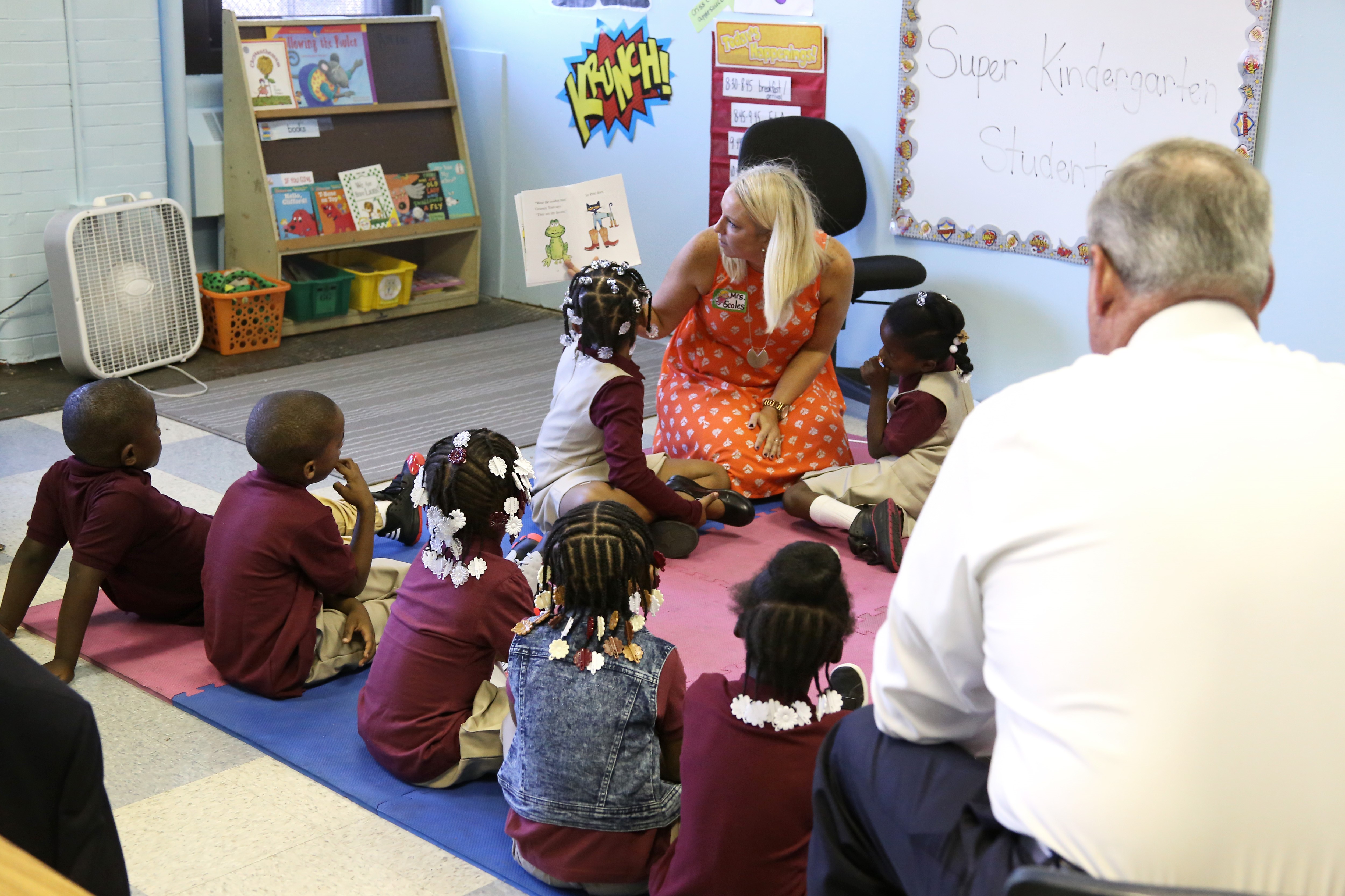 Children sitting in a semi-circle around a teacher holding a book as the mayor watches.